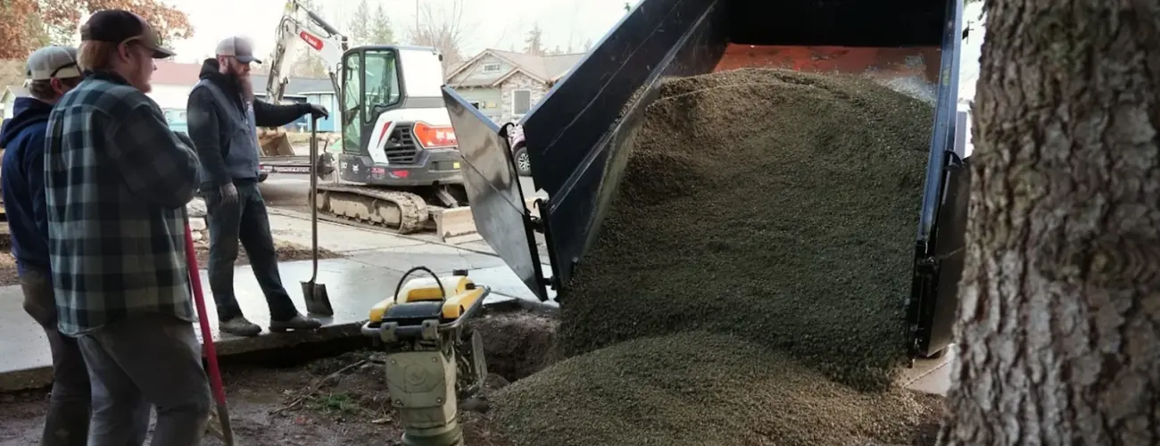 Crew backfilling a trench with gravel beside a mini excavator